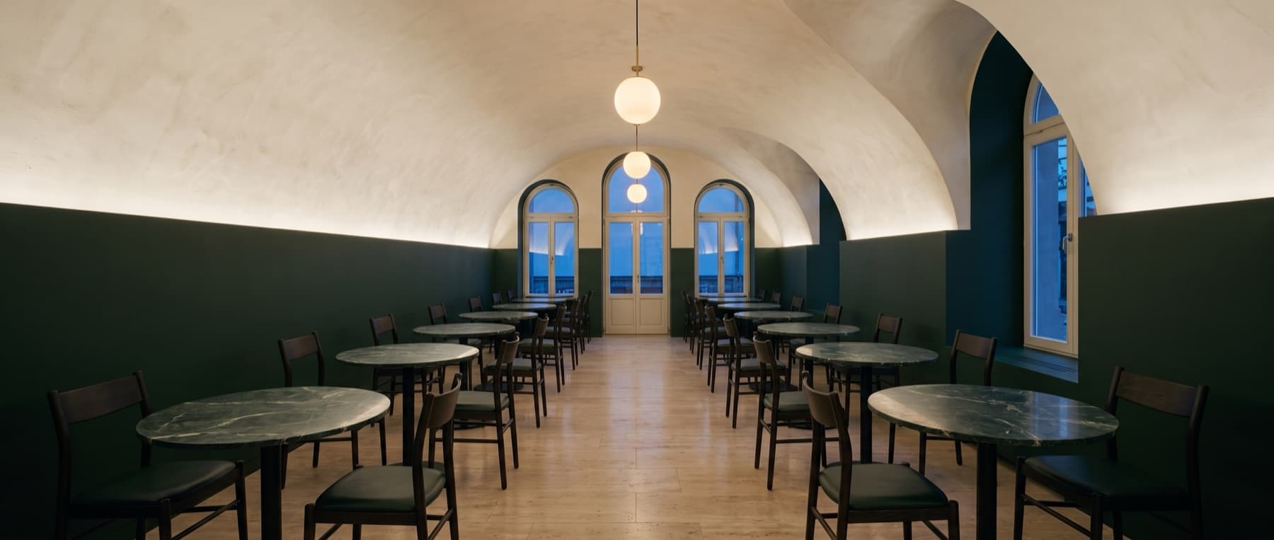 Long vaulted restaurant with green lower walls, rows of round stone tables and dark dining chairs
