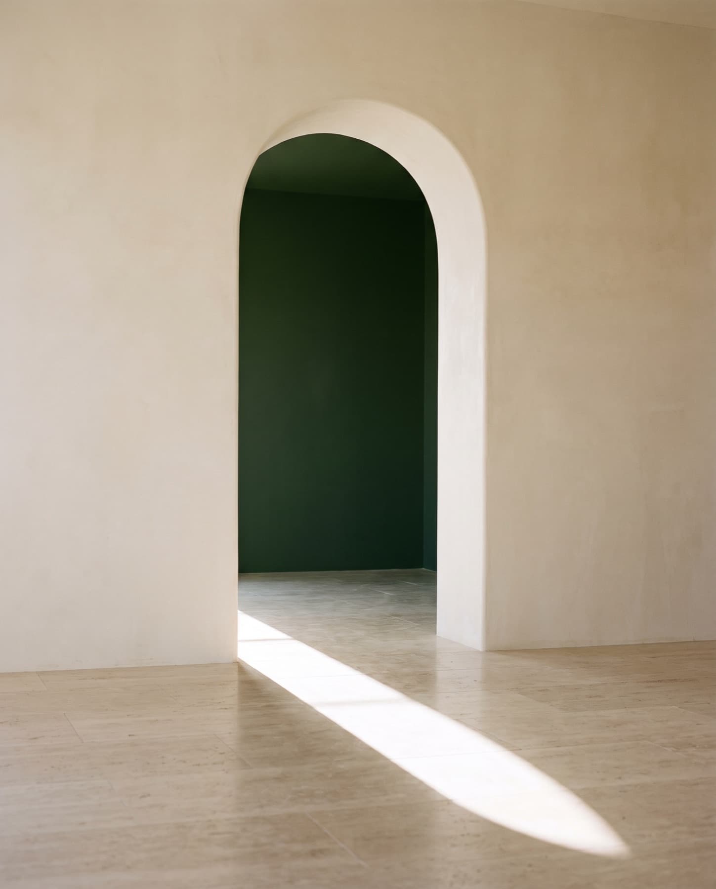 Plastered arched doorway opening onto a dark green room, with sunlight across a stone floor